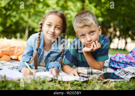 Two smiling kids chilling after school on grass in park while doing homework together outside Stock Photo