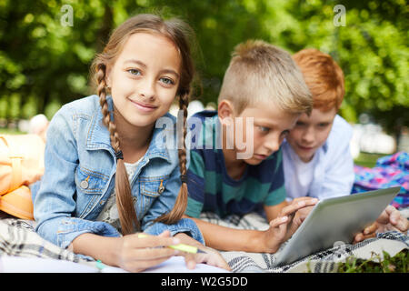 Three school friends spending their summer holidays in park, lying on grass, drawing and playing Stock Photo