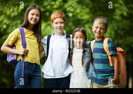 Group of happy interracial friends with satchels standing in park and embracing each other while posing for camera Stock Photo