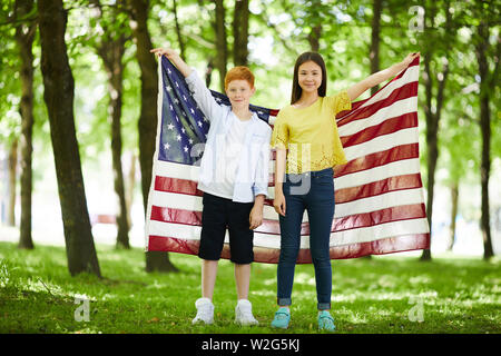 Content multi-ethnic friends in casual outfits standing in forest and raising American flag up Stock Photo