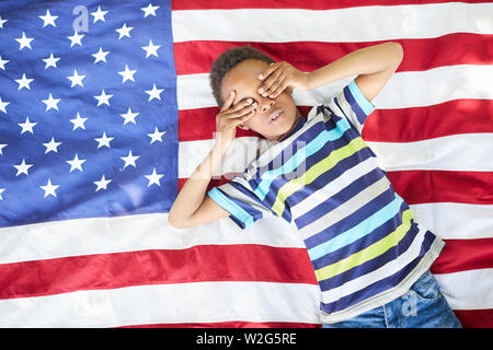 Serious Black boy in stripped tshirt lying on American flag plaid and covering eyes with hands Stock Photo