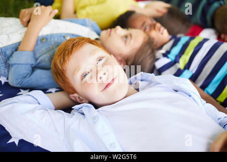 Close-up of pensive dreamy American redhead boy lying on national flag plaid together with multi-ethnic kids Stock Photo