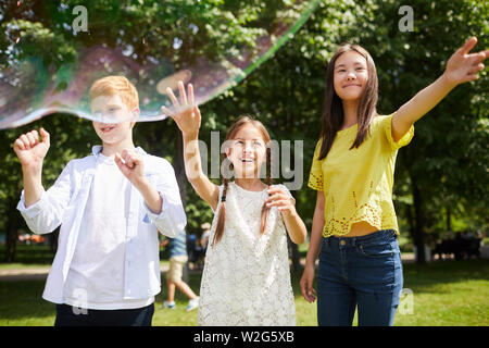 Cheerful carefree friends in casual clothing standing in city park and gesturing hands while looking at soap bubbles Stock Photo