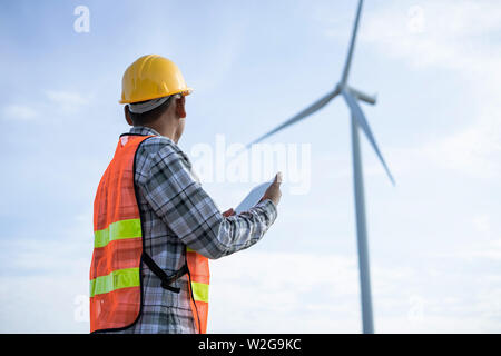 Male Engineer holding tablet checklist in Wind Mill Power Generator Station with orange safety hat and wind turbines on background. Stock Photo