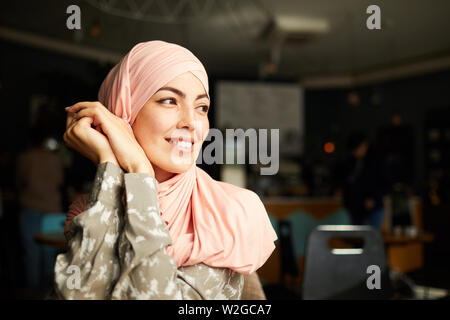 Pretty smiling young Muslim woman sitting at cafe table and looking through window Stock Photo