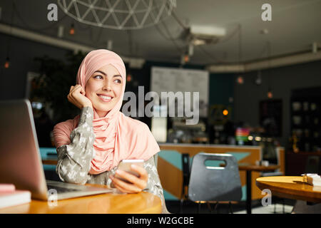 Pretty dreamy young Muslim woman sitting at cafe table with smartphone, smiling and looking through window Stock Photo