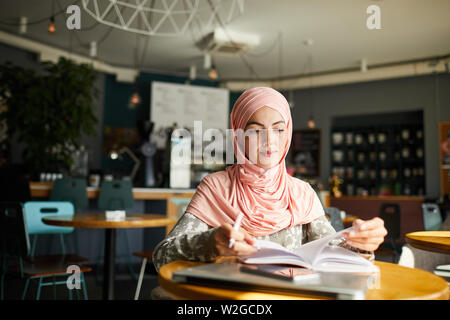 Serious young Muslim woman sitting at cafe table, reading book and taking notes Stock Photo