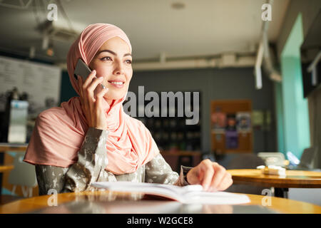 Pretty smiling Muslim businesswoman working at cafe table and making phone call Stock Photo