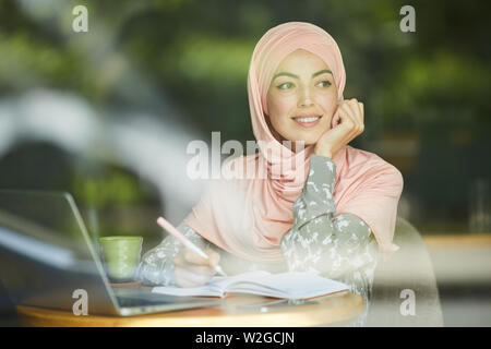 Pretty smiling pensive Muslim female student sitting at cafe table and writing in textbook Stock Photo