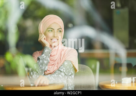 Happy young Muslim businesswoman in traditional dress and hijab sitting at table in restaurant with laptop Stock Photo