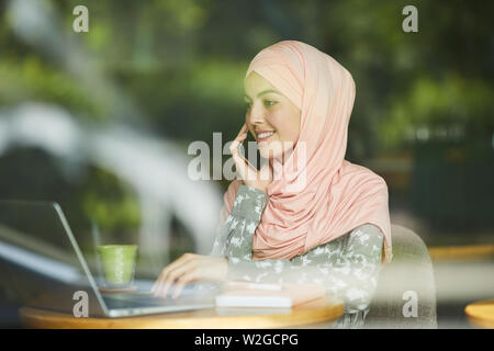 Pretty smiling young Muslim businesswoman in hijab working on laptop and taking on phone Stock Photo