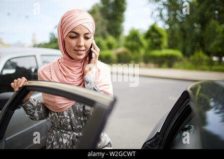 Positive pretty Muslim young woman opening car door and calling on phone Stock Photo