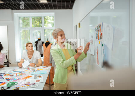 In a studio. Side view of a serious aged blonde woman standing in a studio and pinning a sketch on the whiteboard Stock Photo