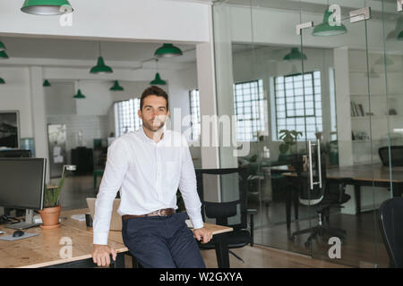 Confident young businessman leaning against an office desk Stock Photo