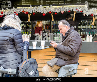 Budapest, Hungary - December 31, 2018: Elderly man uses a mobile phone sitting at table in street cafe with cup of mulled wine. Annual Christmas fair Stock Photo