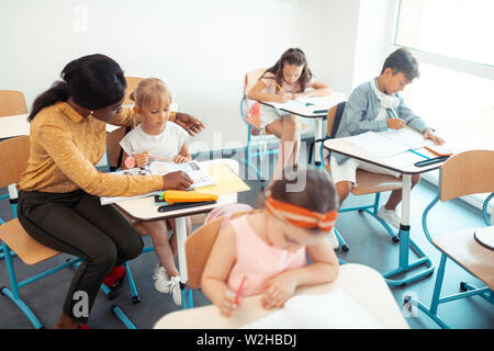 Dark-haired teacher sitting near her pupils helping them with task Stock Photo