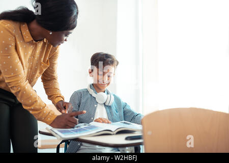 Dark-skinned teacher wearing spotted blouse standing near pupil Stock Photo