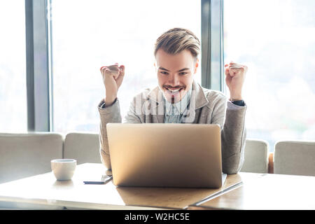 Portrait of handsome amazed satisfied positive bearded young freelancer in grey blazer are sitting in cafe, watching online football match with toothy Stock Photo