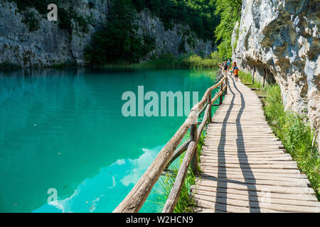 Tourists walking along a boardwalk next to a stunning turquoise colored lake where fish swimming in the crystal clear water - Plitvice Lakes N. P. Stock Photo