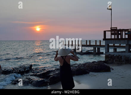 Young attractive woman at the seashore in Vietnam wearing a typical hat staring at the sunset. Young lady watching sunset over the South China Sea in the pier.