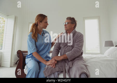 Female doctor consoling active senior man on bed in bedroom at comfortable home Stock Photo