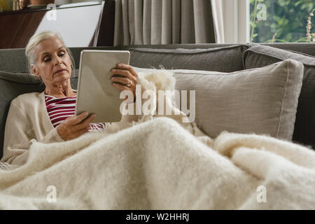 Active senior woman relaxing on sofa and using digital tablet in living room Stock Photo