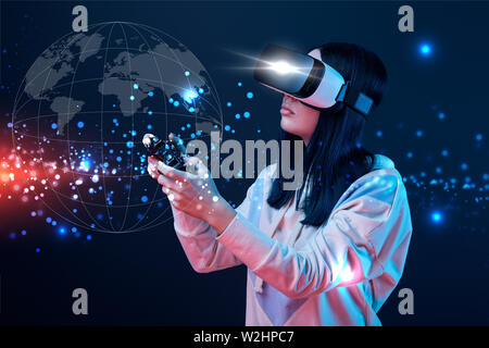 KYIV, UKRAINE - APRIL 5, 2019: Young woman in virtual reality headset using joystick on dark background with globe illustration Stock Photo