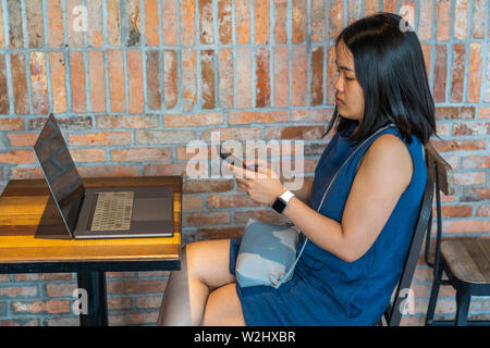 Asian young woman using cellphone next to laptop on table Stock Photo