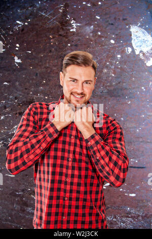 Young man wearing a colorful red checked shirt holding his clenched fists to his chest as he smiles at the camera over a splattered grunge grey backgr Stock Photo