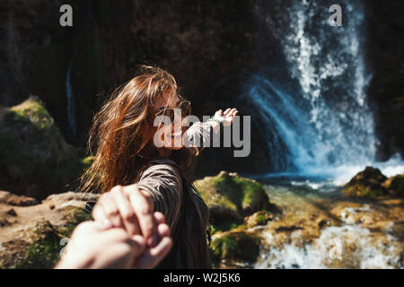 Two hikers in nature are holding hands while crossing the mountain creek. They are enjoying landscape at the beautiful waterfall in mountains. Stock Photo