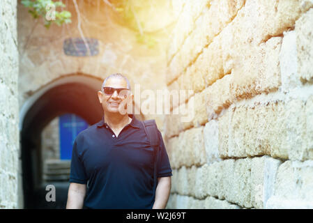 Smiling trendy tourist senior with black t-shirt and sunglasses standing by old ancient city street with brick historic stone wall. Stock Photo