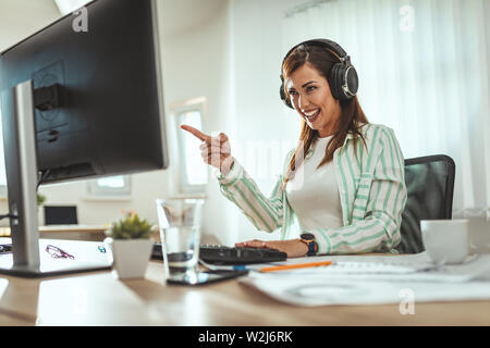 A successful female entrepreneur, with headphones on her head, is listening music on a smartphone and  working on the computer in the office. Stock Photo
