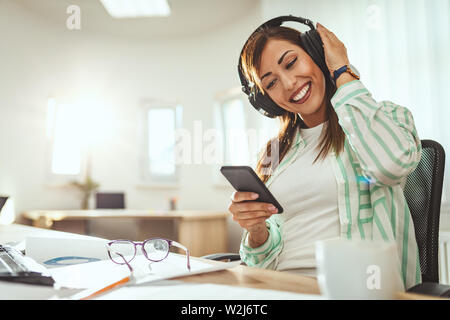 A successful female entrepreneur, with headphones on her head, is searching music on a smartphone in the office. Stock Photo