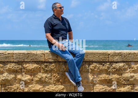 Smiling trendy tourist elderly man sitting by old brick historic stone. Portrait of handsome aged fashion senior wearing black t-shirt and jeans on bl Stock Photo