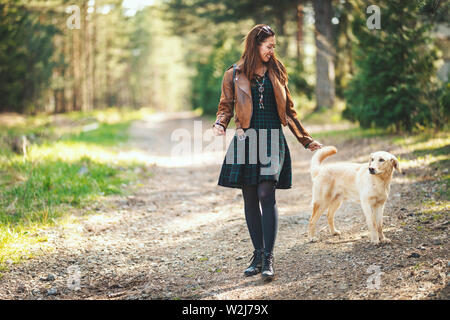 Happy smiling woman is walking along forest path with her dear pet dog and enjoying landscape in forest. Stock Photo
