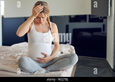 Troubled heavily pregnant young woman sitting cross legged on a bed cradling her swollen abdomen and clutching her head looking down with a serious ex Stock Photo