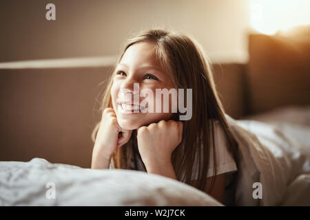 A happy little girl lies on the bed in the morning and laughs after awakening. Stock Photo