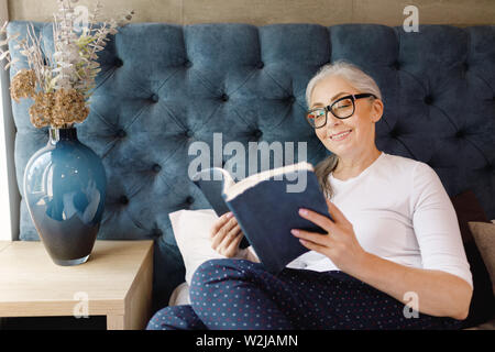 Smiling senior woman with grey hair in eyeglasses laying on the bed and reading book in bedroom Stock Photo