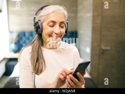 Senior woman with white hair in headphones turn music on the smartphone Stock Photo