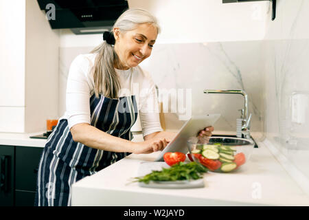 Smiling senior woman with white hair in in an apron using tablet while cooking at the kitchen Stock Photo