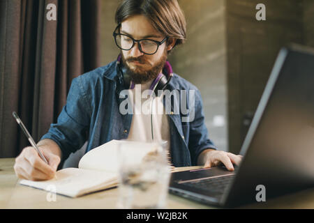 Bearded student man in headphones wearing glasses studying with laptop while writing at the notebook in the evening Stock Photo