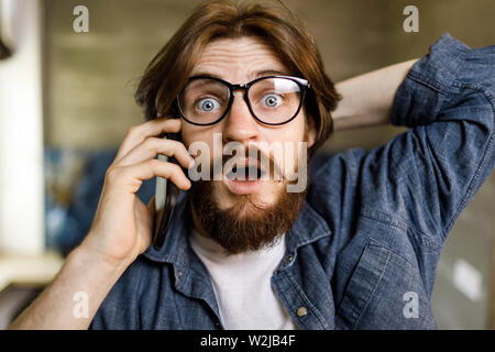 Portrait of shocked bearded hipster man wearing glasses speaking on the smartphone surprised face Stock Photo