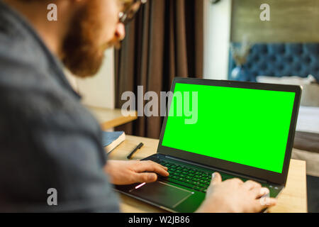 Beardedman typing on laptop with green chroma key screen, sitting indoors, view over shoulder Stock Photo