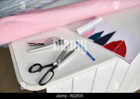 Black scissors, stapler, blue pen, notebook with multicolored textile pieces are lying on the white table indoors. There is a pink fabric roll and a t Stock Photo