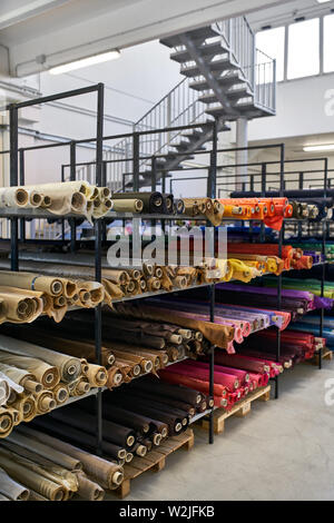 Bright multicolored fabric rolls are lying on the shelves on the stairs background inside a large warehouse. They are brown, white, beige, red, pink, Stock Photo