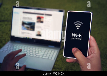 Smartphone with wifi connection generic icon on the screen hold by man on the background of laptop display and keyboard. Man uses Wi-Fi hot spot to Stock Photo