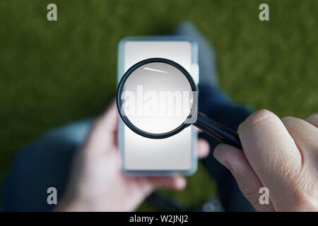 Close up of male hands holding smartphone with magnifying glass above the device screen. Shot of a sitting person with mobile in one hand and the Stock Photo