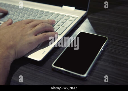 Man sitting behind a laptop and smartphone holding hands on keyboard. Black-screened phone is on table near the notebook with male hands on keypad Stock Photo