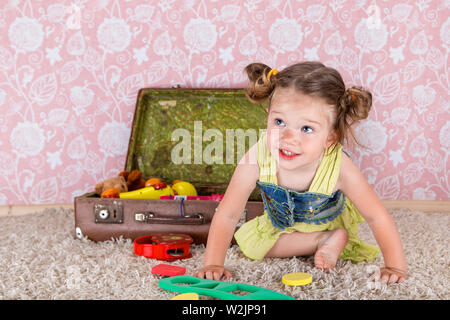 little girl smiling, sitting on the floor in the studio, behind her suitcase with toys. Stock Photo