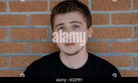 A pale skinned young man with blue eyes and a black shirt with a serious look on his face into the camera leans up against a faded red brick wall. Stock Photo